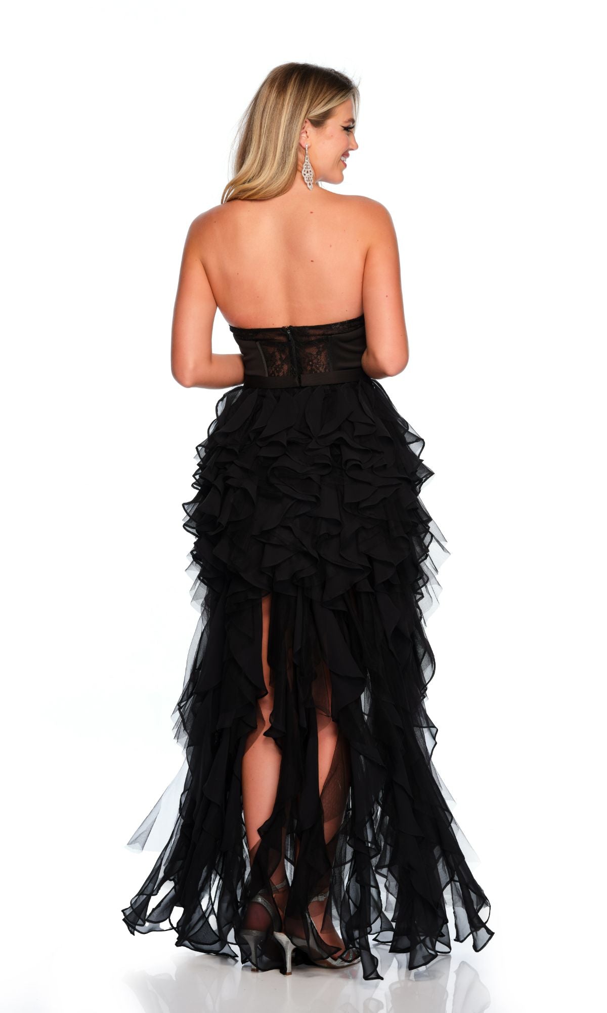 Woman wearing the Long Formal Dress 11446 by Dave and Johnny, a black strapless gown with ruffle layers and a sheer lace back panel, standing and facing away against a white background.